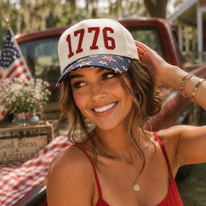 May include: Cream-colored baseball cap with the number "1776" in red lettering. The brim features a blue fabric with red stars. The cap is worn by a person in a red top, with an American flag and a wooden crate in the background.