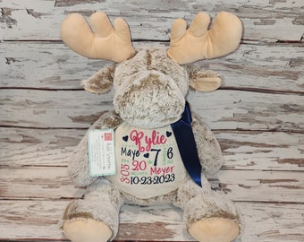 May include: A plush moose toy with embroidered details. The moose has large antlers, a blue ribbon, and a cream-colored patch with the name "Rylie", birth details: 7 lbs, 20 inches, and the date 10-23-2023.