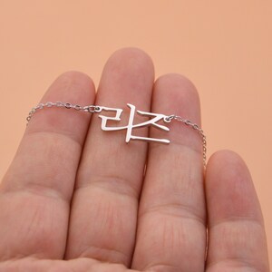 Pu&ograve; includere: Una collana d'argento con una catena delicata e un ciondolo con la parola coreana "라온" in un carattere semplice e moderno.