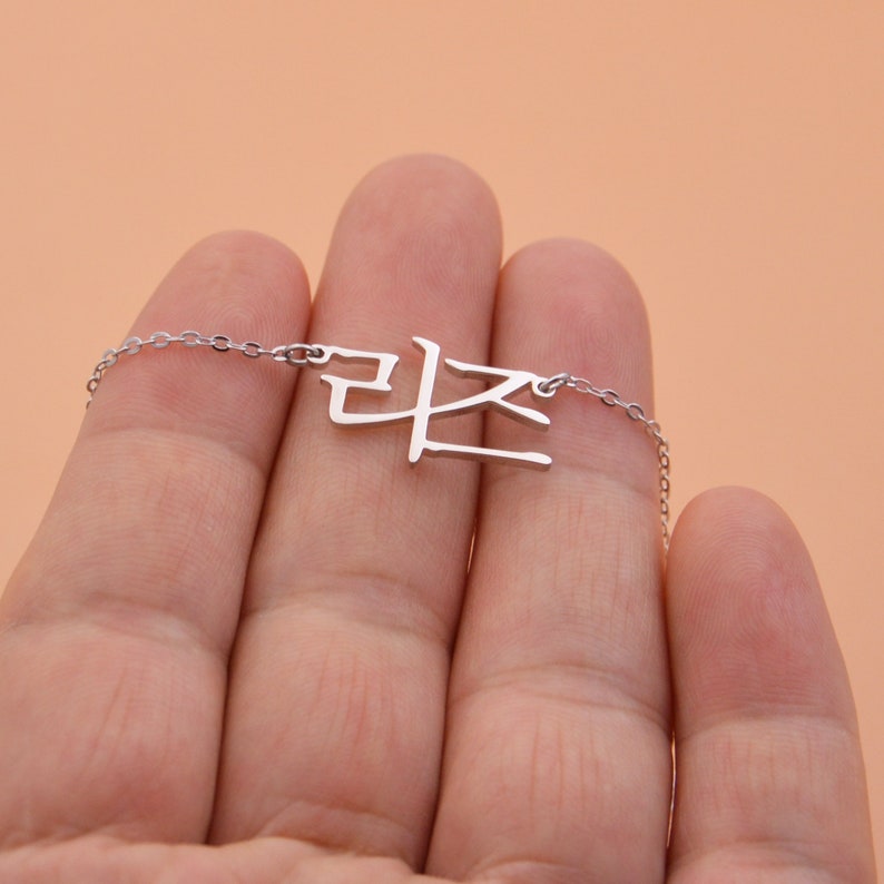 Pu&ograve; includere: Una collana d'argento con una catena delicata e un ciondolo con la parola coreana "라온" in un carattere semplice e moderno.