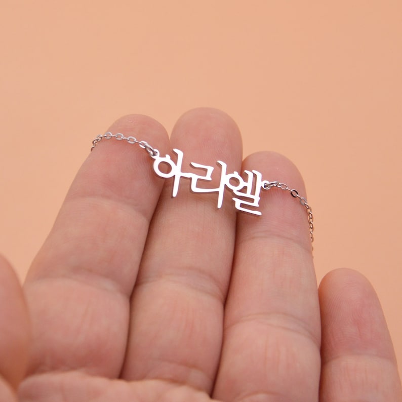 Pu&ograve; includere: Una collana d'argento con un ciondolo a forma di parola coreana che si legge "하리엘".