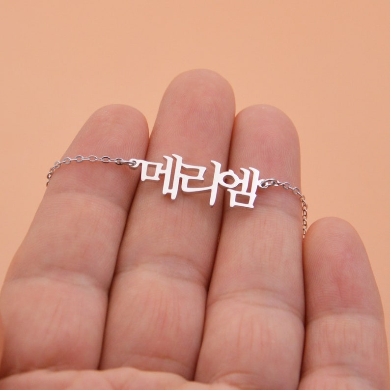 Pu&ograve; includere: Una collana d'argento con un ciondolo a forma di parola coreana che si legge "대한민국".