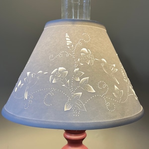 May include: A gray lampshade with a white floral and hummingbird design. The lampshade is made of a fabric-like material and has a scalloped edge.