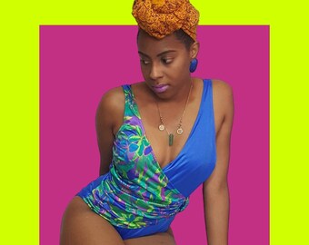 Pode incluir: Uma mulher vestindo um maiô azul e verde com estampa floral e um detalhe de drapeado azul e roxo. Ela está usando um colar de ouro com um pingente de pedra verde e uma faixa de cabeça marrom.