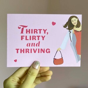Pu&ograve; includere: Carta rosa con il testo "Thirty, flirty and thriving" in lettere rosse. Una donna con lunghi capelli castani &egrave; illustrata con una giacca bianca e un vestito rosso. Tiene una borsa a mano rossa.