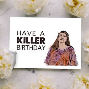 May include: White birthday card with the text "HAVE A KILLER BIRTHDAY" and an illustration of a woman in a colorful patterned top. The card is surrounded by white flowers on a marble surface.