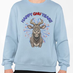 Puede incluir: Sudadera azul claro con un dibujo animado de ñu y el texto "HAPPY GNU YEAR!!" en azul y rojo. El ñu tiene cuernos grandes y una amplia sonrisa, con fuegos artificiales en el fondo.