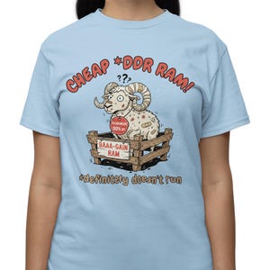 May include: Light blue t-shirt with the text "CHEAP *DDR RAM!" in red and a cartoon ram in a wooden crate. The ram has bandages and a sign that says "BAAA-GAIN RAM". Below the crate is the text "*definitely doesn't run".