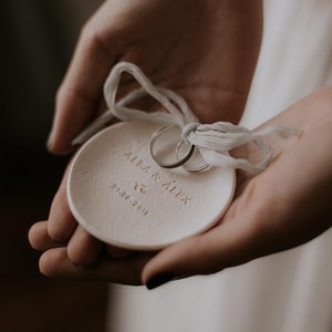 May include: White ceramic ring dish inscribed with "Alba & Alex" and the date "21.07.2018" in gold lettering. Two gold wedding bands are placed on the dish.