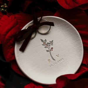 May include: A round white ceramic ring dish featuring a stamped holly sprig illustration with red berries, the letters R and V, and the date 07-12-2024. A dark red velvet ribbon is tied around two gold-toned metal rings resting on the dish surface.