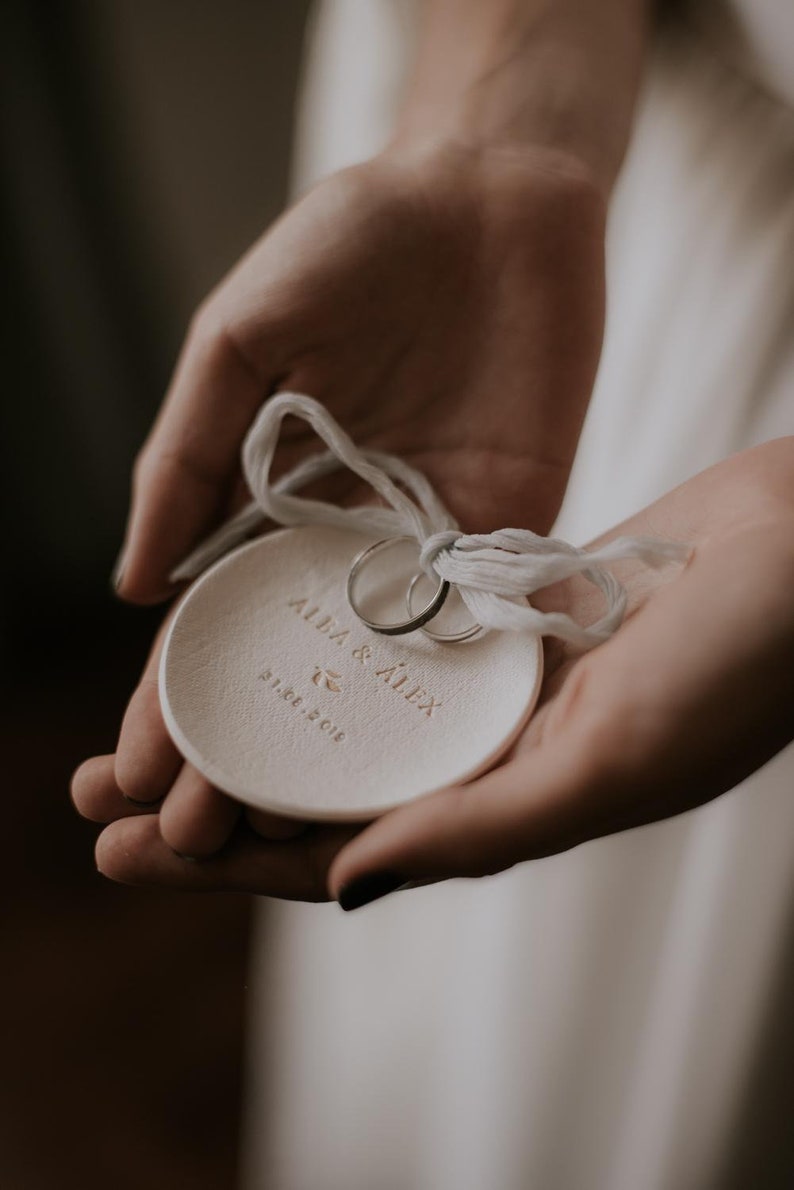 May include: White ceramic ring dish inscribed with "Alba & Alex" and the date "21.07.2018" in gold lettering. Two gold wedding bands are placed on the dish.