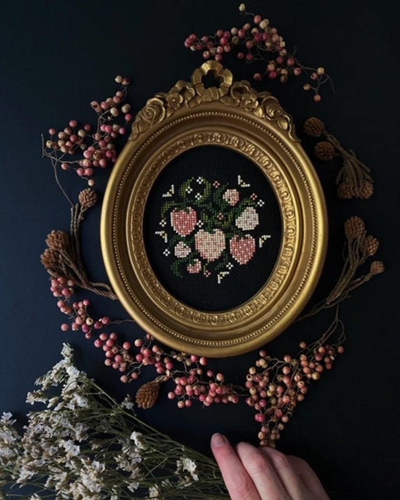 May include: A framed cross-stitch embroidery featuring a bouquet of pink and white flowers with green leaves on a black background. The frame is gold and ornate.