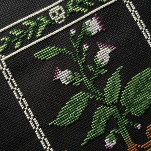 May include: A black and white cross-stitch embroidery of a plant with green leaves and white flowers in a brown pot. The plant is surrounded by a white border.