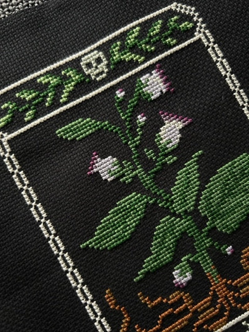May include: A black and white cross-stitch embroidery of a plant with green leaves and white flowers in a brown pot. The plant is surrounded by a white border.