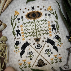 May include: A cross-stitch embroidery pattern featuring a black crow, a sunflower, a beehive, and other floral elements. The design is stitched on a white fabric with black, yellow, and green thread.