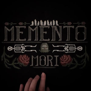Può includere: Tessuto nero con un disegno a punto croce che presenta le parole "Memento Mori" in filo bianco. Il disegno include anche due rose rosse, un teschio e uno scheletro.