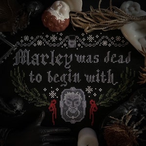 May include: Black and white cross-stitch embroidery with the text "Marley was dead to begin with". The embroidery is framed with a black border and features a small image of a ghost in the center.