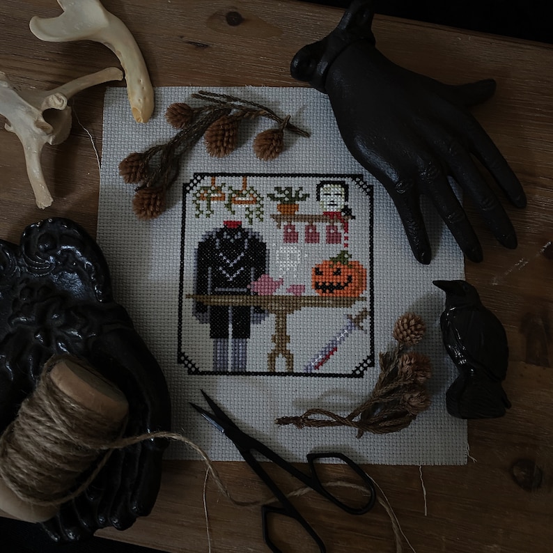 Op de afbeelding: Een kruissteekpatroon met een koploze figuur in een zwart pak, een pompoen en een mes. Het patroon is omlijst door een witte rand en wordt weergegeven op een houten oppervlak.