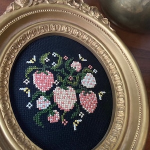May include: A gold oval frame with a black background and a cross-stitch design of pink and white flowers with green leaves.