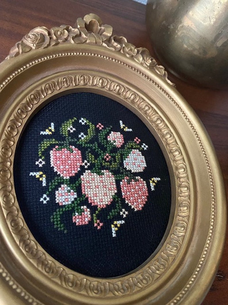 May include: A gold oval frame with a black background and a cross-stitch design of pink and white flowers with green leaves.