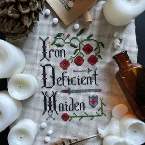 May include: Cross-stitch embroidery with the text "Iron Deficient Maiden" in a gothic font. The embroidery is surrounded by white candles, a small bottle, and a feather.