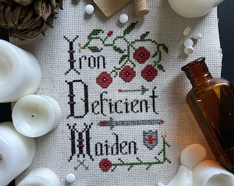 May include: Cross-stitch embroidery with the text "Iron Deficient Maiden" in a gothic font. The embroidery is surrounded by white candles, a small bottle, and a feather.