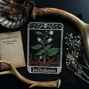 Könnte beinhalten: Ein schwarzes und weißes Kreuzstichmuster einer Tollkirschenpflanze mit grünen Blättern und weißen Blüten. Das Muster ist in einem braunen und weißen Rahmen eingefasst und das Wort "Belladonna" ist unter der Pflanze gestickt. Das Muster wird auf einem schwarzen Stoffhintergrund mit Geweih und einem Buch mit dem Titel "The Dark Ages" im Hintergrund angezeigt.