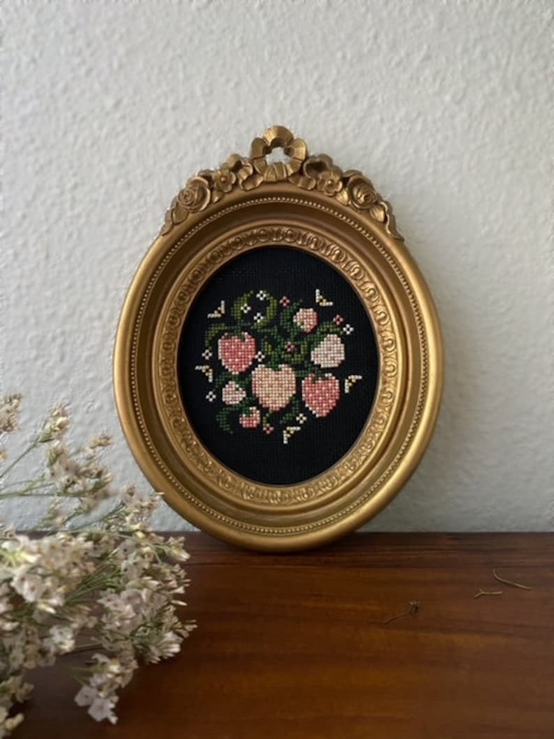 May include: A gold framed picture with a black background and a cross-stitch design of pink hearts and green leaves.