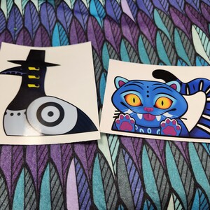 May include: Two stickers on a patterned background. One sticker features a stylized black and gray bird-like figure with a hat. The other sticker is a blue cartoon cat with yellow eyes and a striped tail.