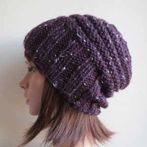 K&ouml;nnte beinhalten: Eine violette Strickm&uuml;tze mit wei&szlig;en Flecken. Die M&uuml;tze ist locker und hat eine gerippte Struktur.