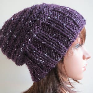 K&ouml;nnte beinhalten: Eine slouchy Beanie M&uuml;tze in einem tiefen Lila mit wei&szlig;en Flecken im gesamten Strick. Die M&uuml;tze ist aus einem weichen, dicken Garn gefertigt und hat eine slouchy Passform.