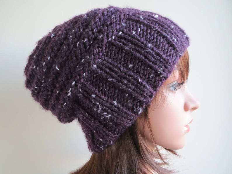 K&ouml;nnte beinhalten: Eine slouchy Beanie M&uuml;tze in einem tiefen Lila mit wei&szlig;en Flecken im gesamten Strick. Die M&uuml;tze ist aus einem weichen, dicken Garn gefertigt und hat eine slouchy Passform.