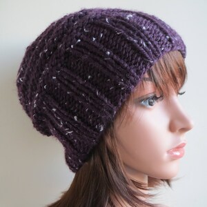 K&ouml;nnte beinhalten: Eine violette Strickm&uuml;tze mit wei&szlig;en Flecken. Die M&uuml;tze wird von einer Puppe mit braunen Haaren getragen.
