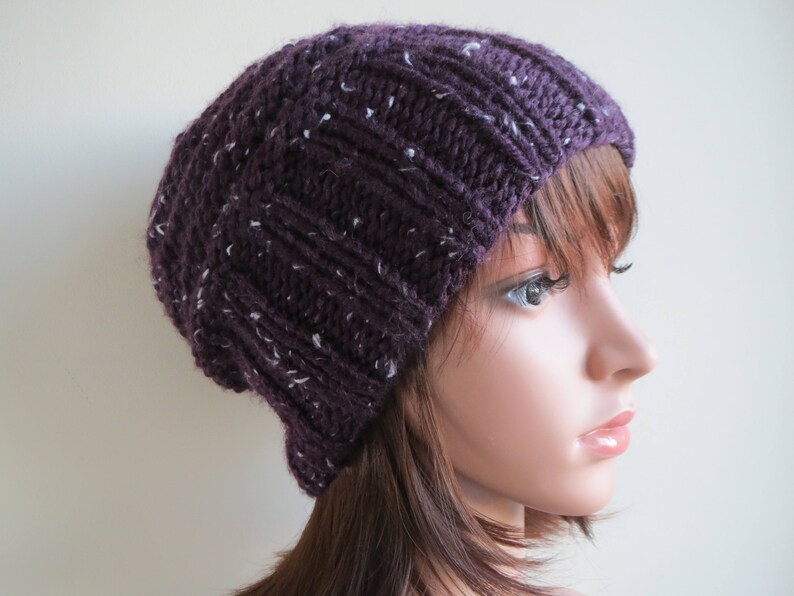K&ouml;nnte beinhalten: Eine violette Strickm&uuml;tze mit wei&szlig;en Flecken. Die M&uuml;tze wird von einer Puppe mit braunen Haaren getragen.