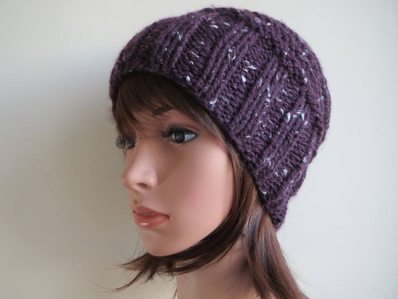 K&ouml;nnte beinhalten: Eine lilafarbene Strickm&uuml;tze mit wei&szlig;en Flecken. Die M&uuml;tze ist aus weichem, strukturiertem Garn gefertigt und hat eine lockere Passform.