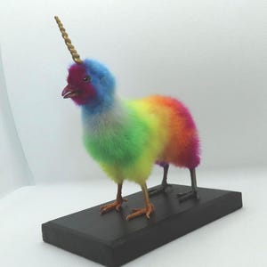 Può includere: Un pollo arcobaleno con un corno di unicorno dorato sulla testa. Il pollo è in piedi su una base nera.