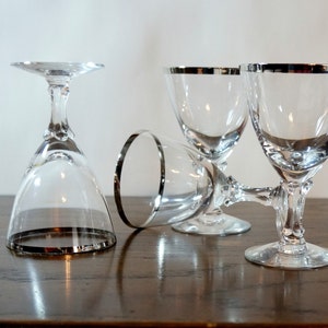 Peut inclure: Quatre gobelets en verre transparent avec des bords et des pieds argentés sont disposés sur une surface en bois sombre. Un gobelet est à l'envers, les autres sont droits ou inclinés, mettant en valeur le design élégant.