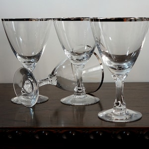 Op de afbeelding: Vier heldere glazen wijnglazen met een zilveren rand. De glazen zijn in een rij op een donker houten oppervlak gerangschikt.