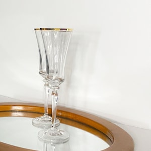 May include: Two clear champagne flutes with gold rims. The flutes have a tall, slender design with vertical ridges. They are placed on a mirrored surface with a wooden border, reflecting the glasses.