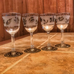 Könnte beinhalten: Vier klare Weingläser aus Glas mit einem matten Design aus silbernen Blättern. Die Gläser sind in einer Reihe auf einer Holzoberfläche angeordnet.