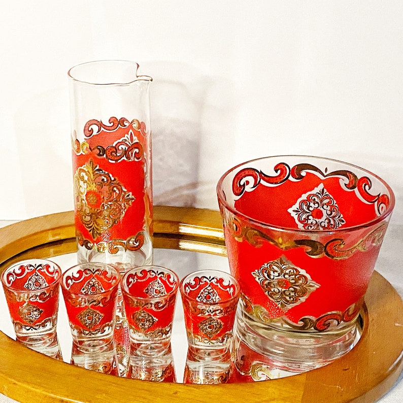 K&ouml;nnte beinhalten: Set aus f&uuml;nf roten und goldenen Gl&auml;sern mit einem dekorativen Muster. Das Set enth&auml;lt einen hohen Krug und vier kleinere Gl&auml;ser. Die Gl&auml;ser sind alle klar mit einem roten und goldenen Design.