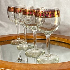 Puede incluir: Cuatro copas de vino de vidrio transparente con un borde dorado y rosa alrededor de la parte superior. Las copas están sobre una superficie de madera con un centro de espejo.