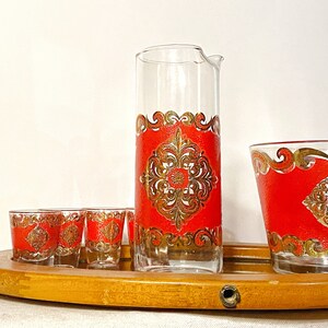 K&ouml;nnte beinhalten: Ein Set aus sechs Vintage-Gl&auml;sern in Rot und Gold mit einem dekorativen Muster. Das Set enth&auml;lt eine Karaffe und f&uuml;nf Schnapsgl&auml;ser. Die Gl&auml;ser stehen auf einem Tablett aus Holz.