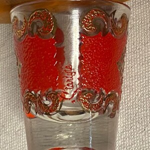 K&ouml;nnte beinhalten: Ein klares Glas-Schnapsglas mit einem roten und goldenen Design. Das Glas hat eine strukturierte Oberfl&auml;che und einen goldenen Rand.