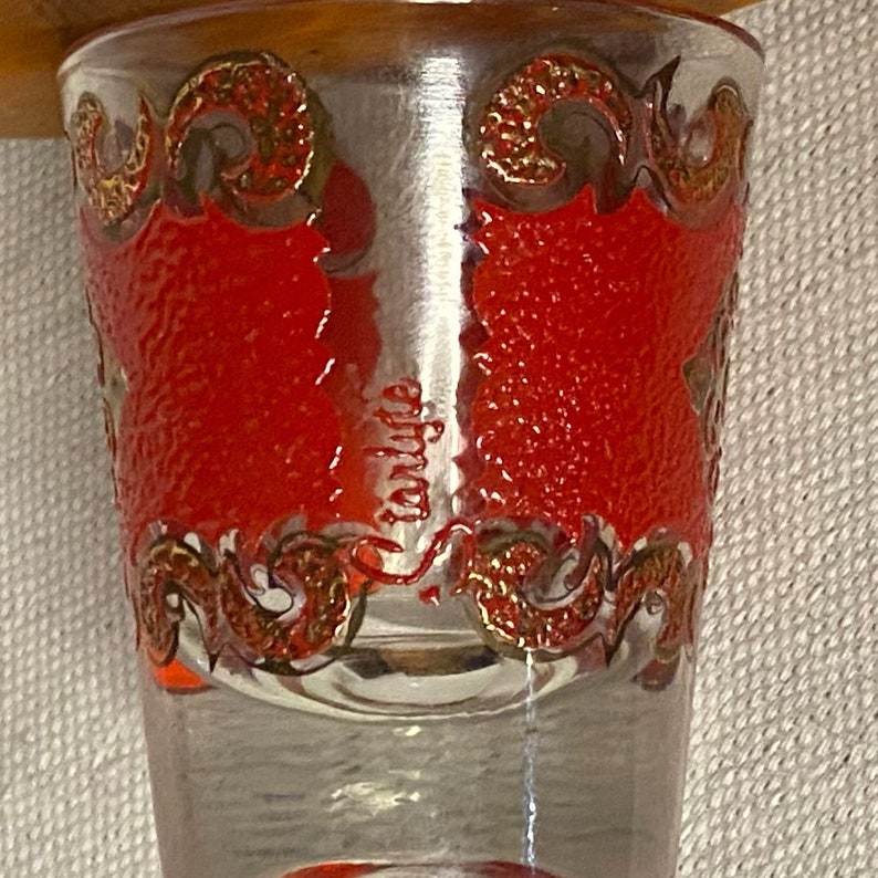 K&ouml;nnte beinhalten: Ein klares Glas-Schnapsglas mit einem roten und goldenen Design. Das Glas hat eine strukturierte Oberfl&auml;che und einen goldenen Rand.