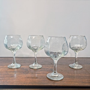 May include: Four clear, textured glass goblets with rounded bowls and slender stems. The glasses are arranged on a dark wooden surface, with a plain white background. The goblets have a unique, textured pattern on the bowl.