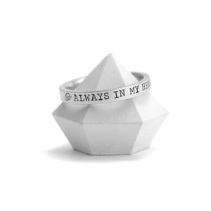 Op de afbeelding: Een zilveren ring met de inscriptie "ALWAYS IN MY HEART" op een witte geometrische standaard.
