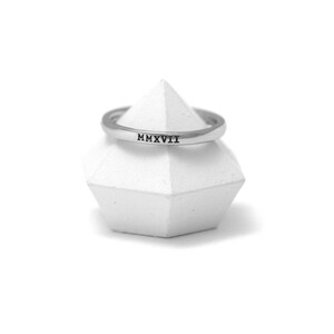 Può includere: Un anello d'argento con l'iscrizione in numeri romani "MMXVII" su un espositore geometrico bianco.