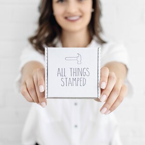 Op de afbeelding: Een witte doos met de tekst "ALL THINGS STAMPED" en een kleine afbeelding van een hamer.