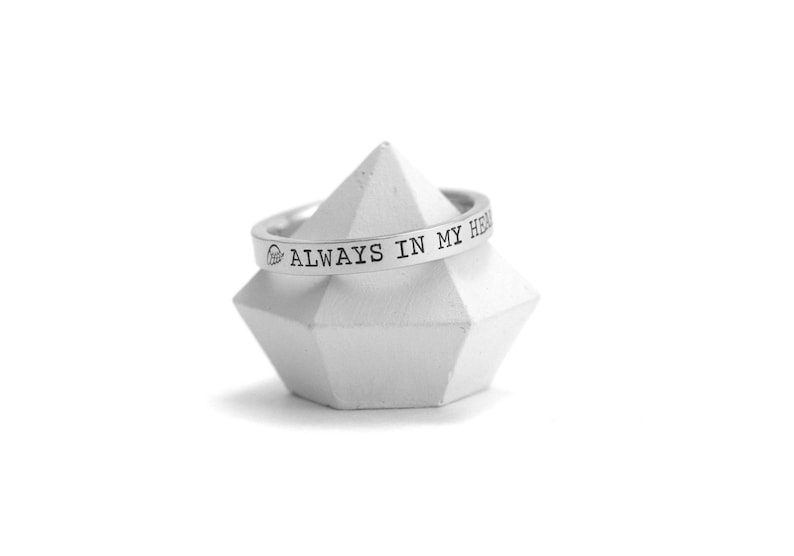 Op de afbeelding: Een zilveren ring met de inscriptie "ALWAYS IN MY HEART" op een witte geometrische standaard.